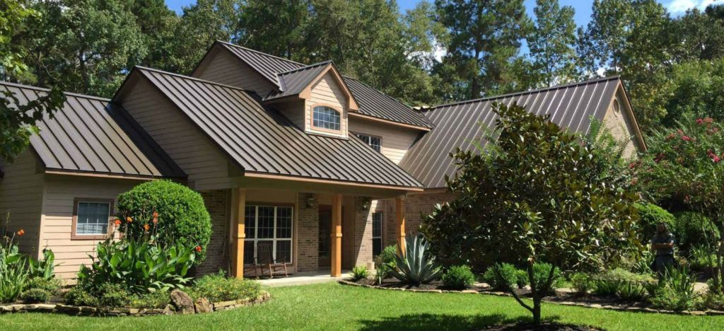 A two-story suburban house with a brown metal roof, tan siding, stone accents, and a lush, landscaped front lawn.