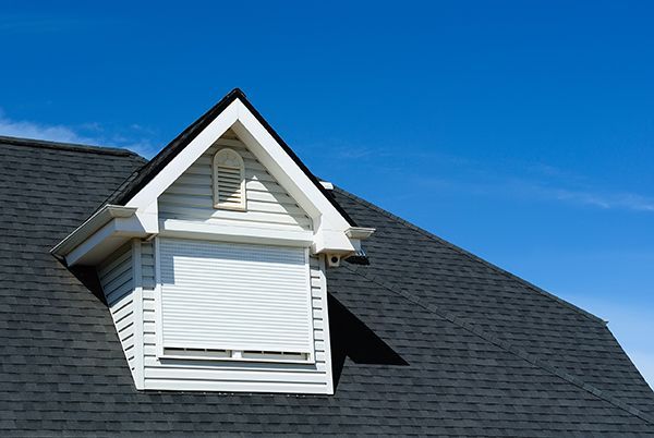 A white dormer window with a closed shutter on a dark shingled roof against a bright blue sky.