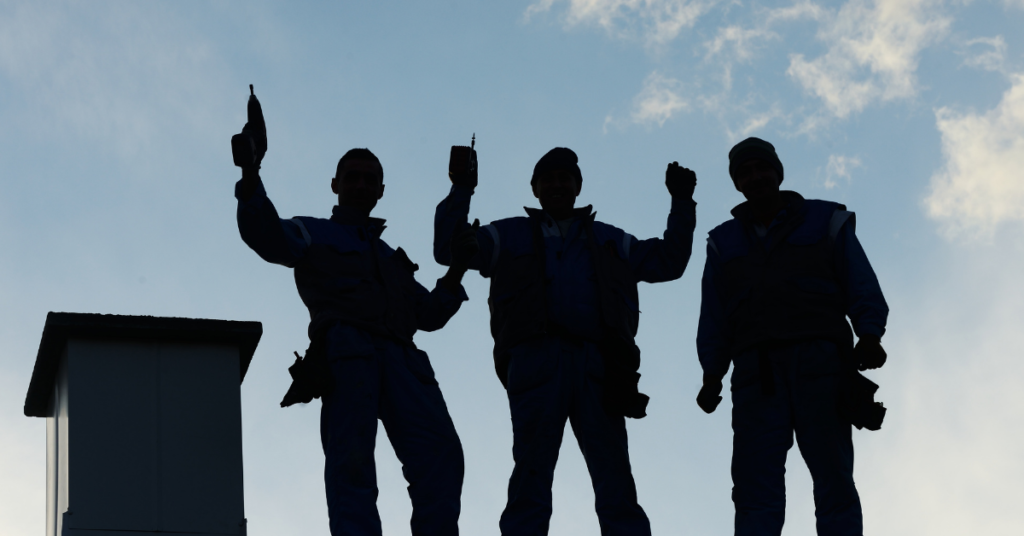 Silhouetted workers standing on a roof against a blue sky, some with arms raised.