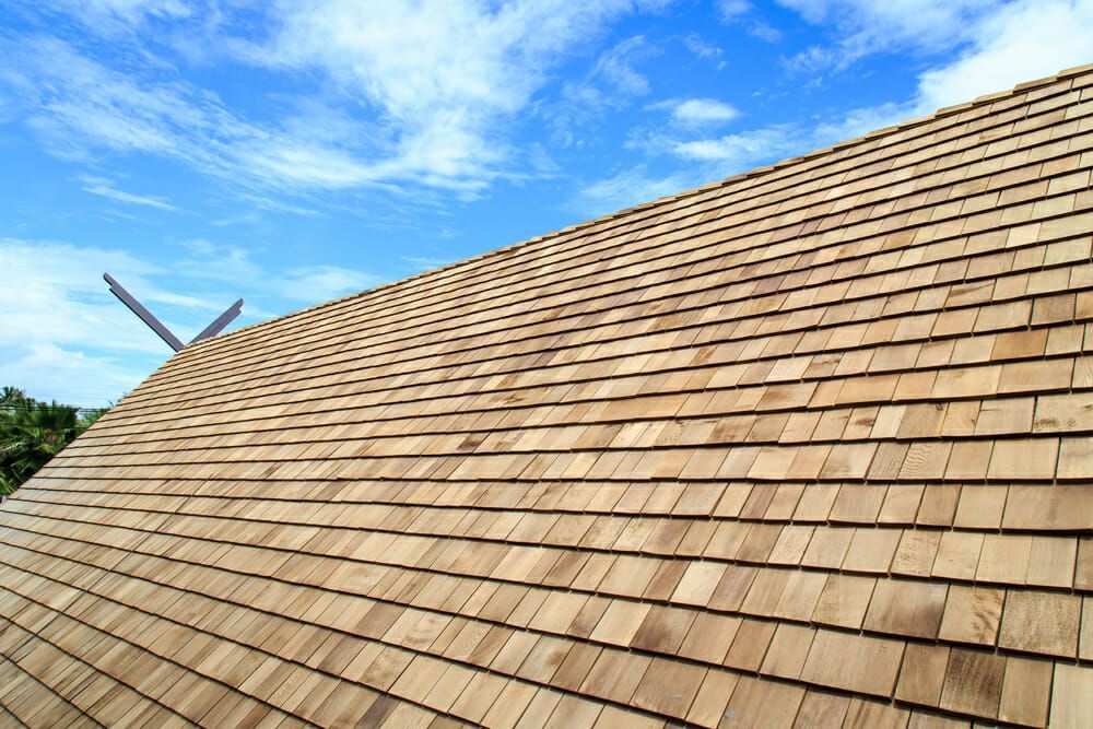 Close-up of a curved roof covered in light-brown wood shingles against a bright blue sky with wispy clouds.