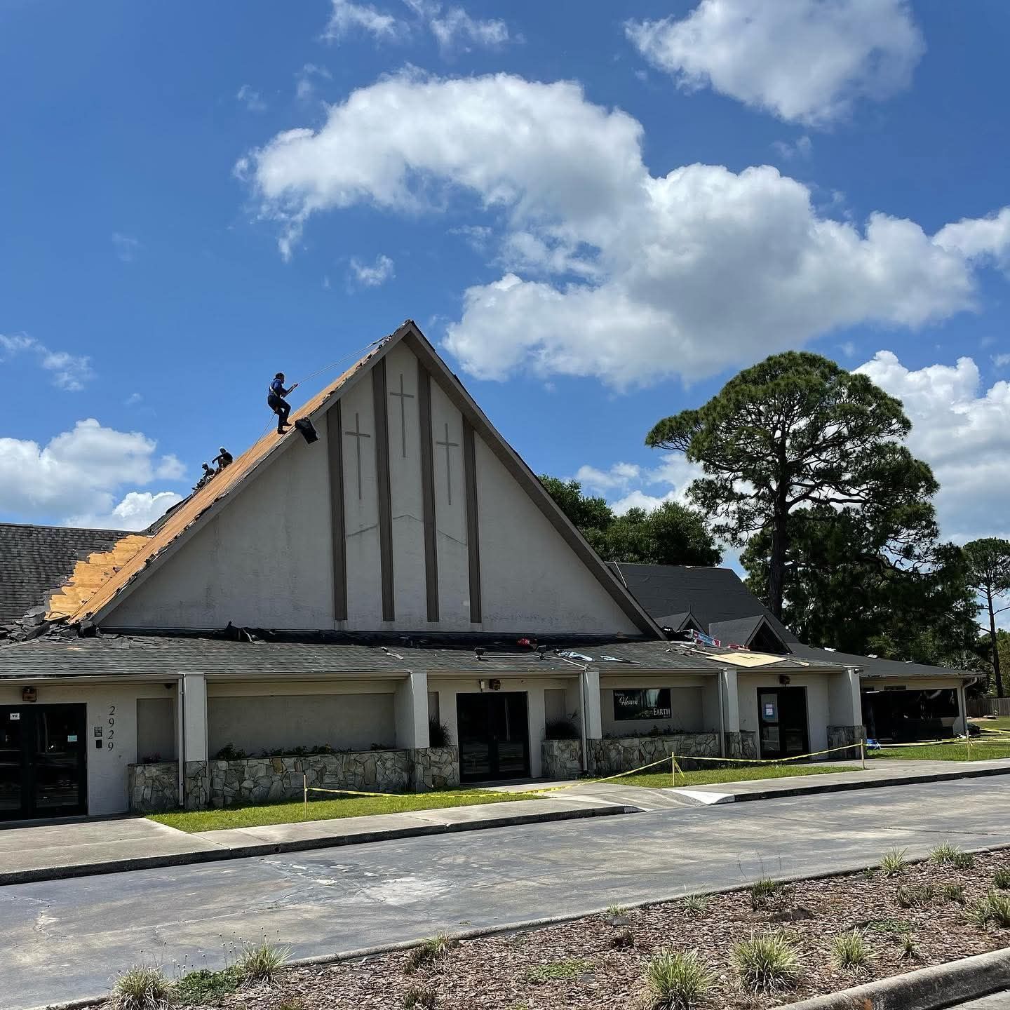 Workers remove shingles from the steep, triangular roof of a light-colored building under a sunny, blue sky.