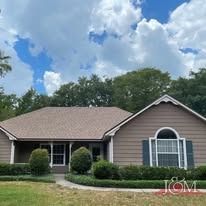 A single-story, taupe-colored suburban house with a brown shingled roof, a front porch, and manicured green bushes.