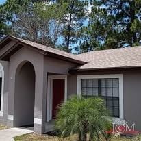 A taupe, single-story house with a red front door, arched entryway, brown roof, and a small palm tree in the front yard.
