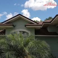 A light green house with a dark brown roof and a large palm tree in the foreground under a blue sky with white clouds.