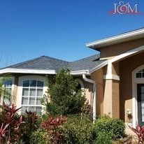 A tan house with a dark gray shingled roof, white trim, and a clear blue sky, partially obscured by green shrubs.