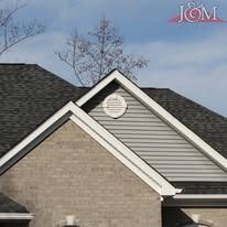 A close-up of a house roof showing dark shingles, a light brown brick exterior, and a white gable vent on light siding.