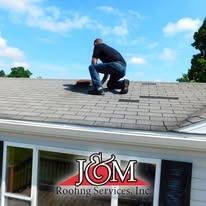 A person kneeling on a shingled roof for repair work, with the J&M Roofing Services, Inc. logo overlaid in the foreground.