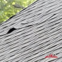 A close-up view of grey asphalt roof shingles, showing one shingle lifting or curling upward.