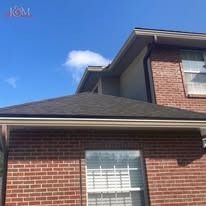 A brick residential home exterior features a dark shingled roof, white gutters, and a window under a clear blue sky.