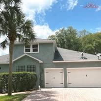 A two-story grey stucco house with a two-car garage and a large palm tree in the front yard under a blue sky.