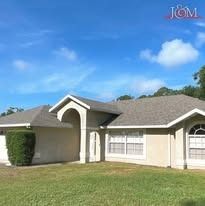 A one-story tan suburban house with a gray shingled roof, a recessed entryway, and a manicured hedge on a grassy lawn.
