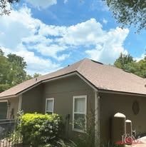 A taupe-colored house with a brown shingled roof under a bright blue sky with scattered white clouds.