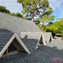 A gray shingled roof with a series of triangular dormers under a clear sky and large green trees.