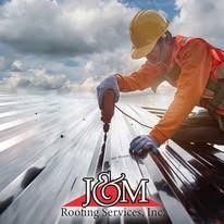 A worker in a yellow hard hat and orange vest uses a drill to install metal roofing. J&M Roofing Services, Inc. logo below.
