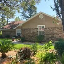 A single-story home with tan horizontal siding, brick exterior, and a gabled roof, surrounded by trees and landscaping.