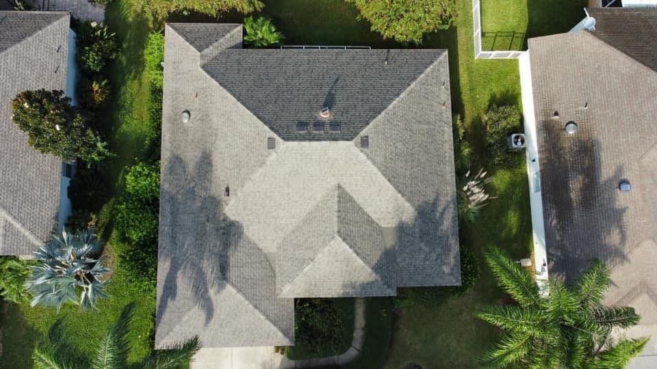 Aerial view of a residential house with a dark shingled roof, surrounded by green trees and palm shadows.
