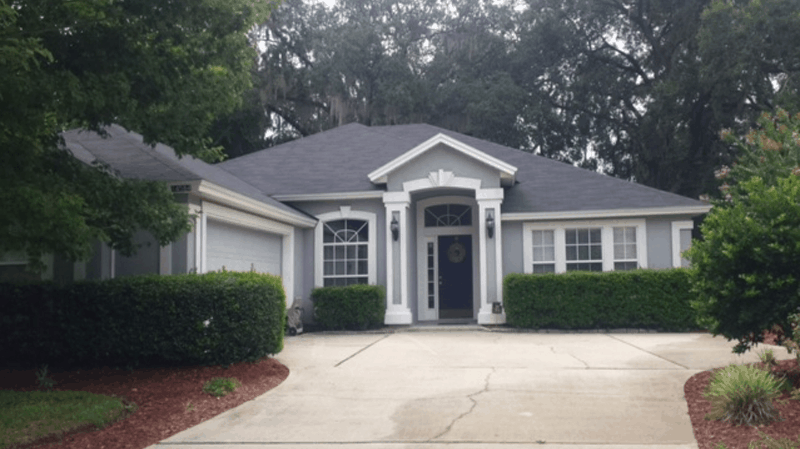 A single-story gray house with white trim and a front-facing garage, surrounded by green hedges and mature trees.