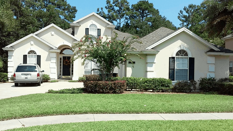 A single-story, cream-colored house with dark shutters, a front yard, a driveway with a silver SUV, and surrounding trees.