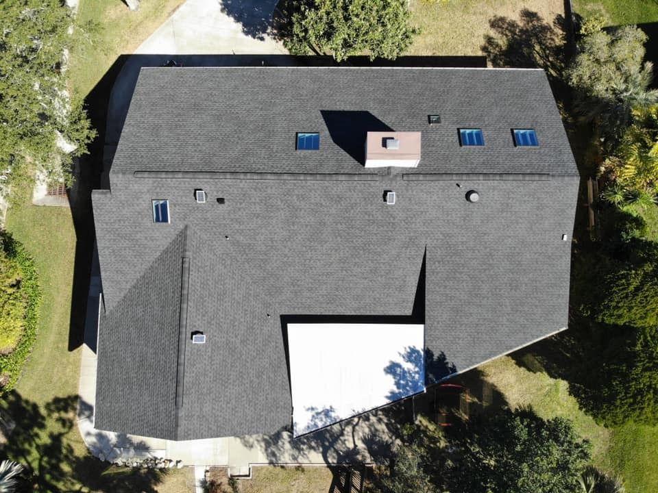 An aerial view of a dark shingled residential roof with a white flat section, a central chimney, and several skylights.