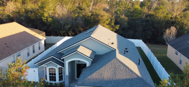 Aerial view of a residential house with a dark gray shingled roof, surrounded by a white fence and lush green trees.
