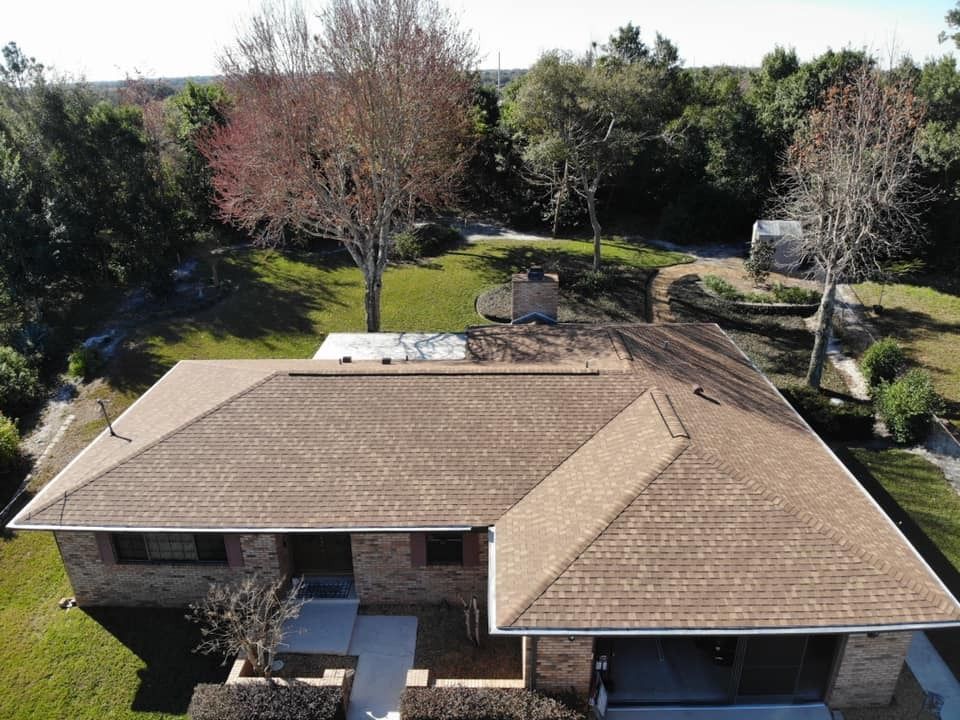 An aerial view of a brick house with a brown shingled roof, surrounded by trees and a lawn.
