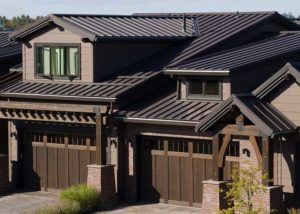 Two-car garage with dark brown paneled doors, brick-base pillars, and a dark standing-seam metal roof with dormer windows.
