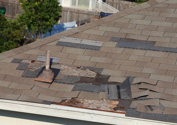Damaged brown asphalt roof shingles with several missing sections, showing underlying material and a vent pipe.