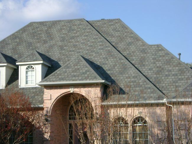A large house with a multi-gabled gray shingle roof, tan brick exterior, and arched entryway under a clear blue sky.