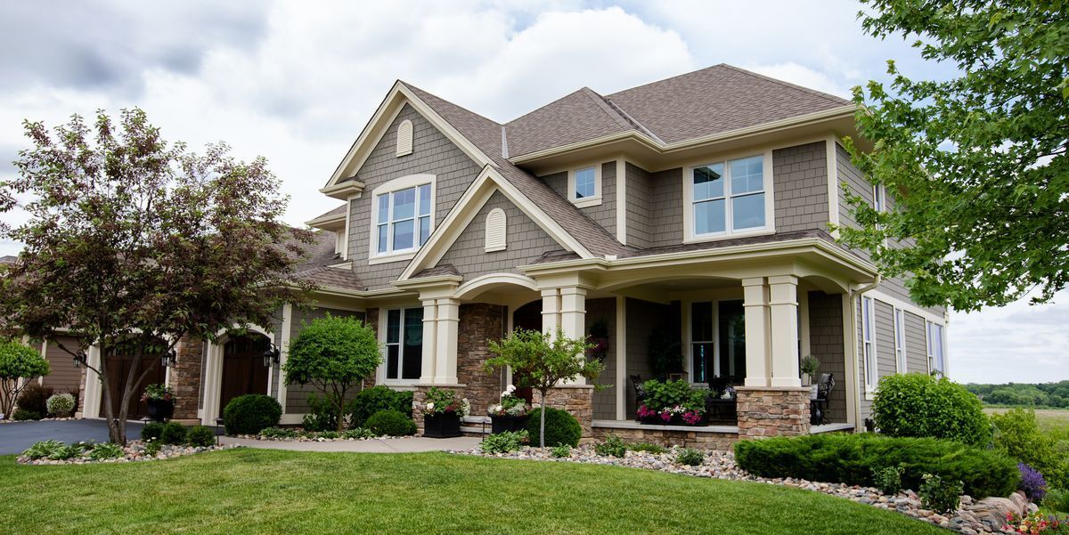 A two-story grey shingled house with a stone porch and pillars, surrounded by a lush green lawn and trees under a cloudy sky.