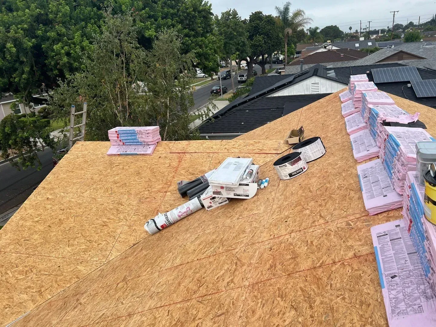Rooftop with OSB decking and pink insulation, with tools and materials staged for installation, trees and neighborhood in the background.