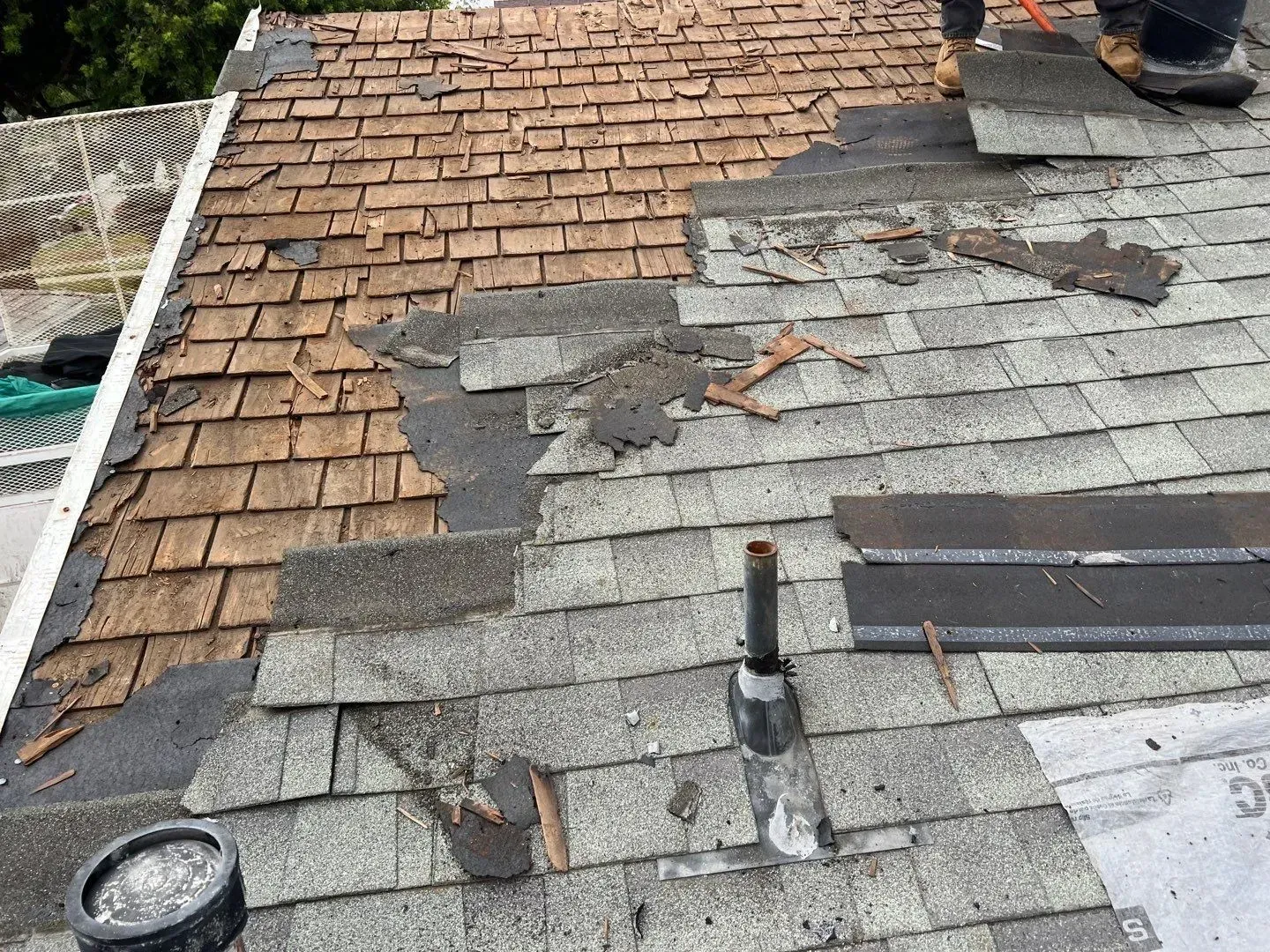 Roof partially stripped of shingles, showing old and new layers with person's legs visible.