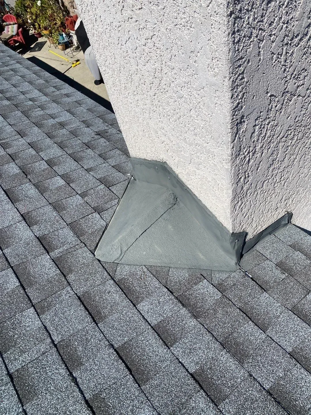 Close-up of a roof with dark gray shingles and a gray stucco chimney. The chimney has flashing at its base.