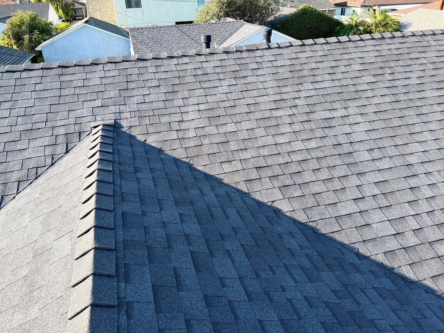 Gray asphalt shingle roof on a house, with a triangular-shaped edge in the foreground and other houses in the background.