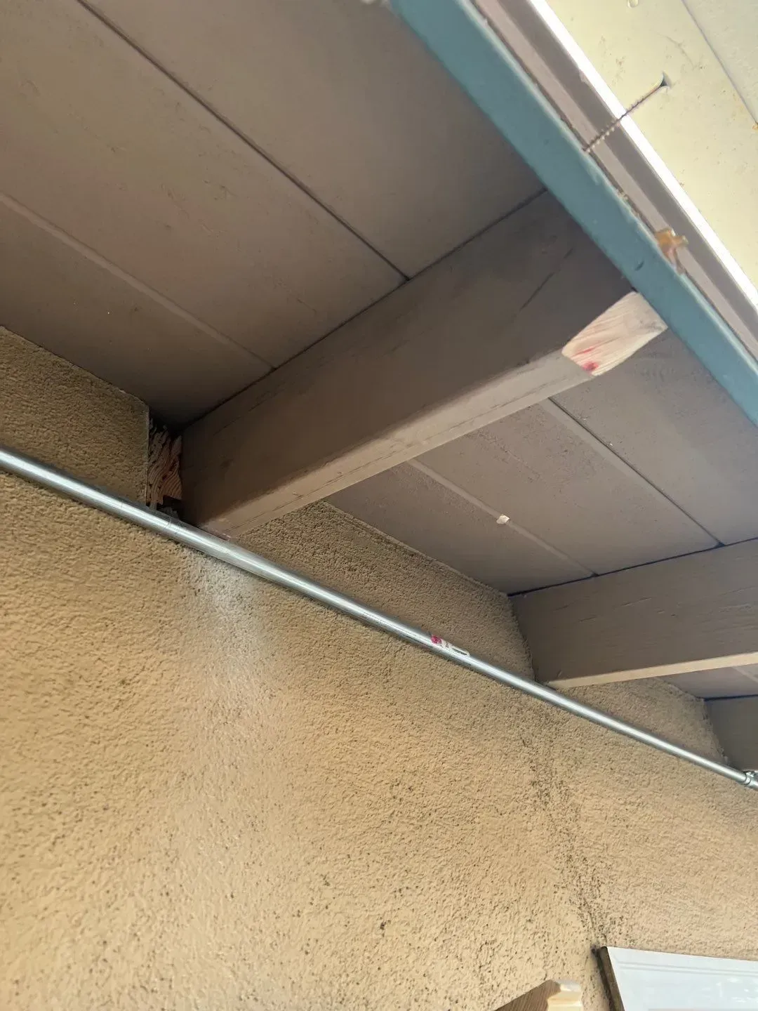Beams and ceiling of a patio with a conduit running along the stucco wall.