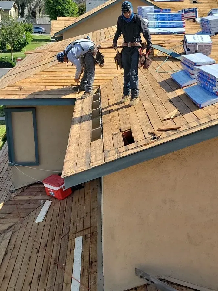 Two roofers installing shingles on a house roof. One bends, the other stands. Blue and brown hues.
