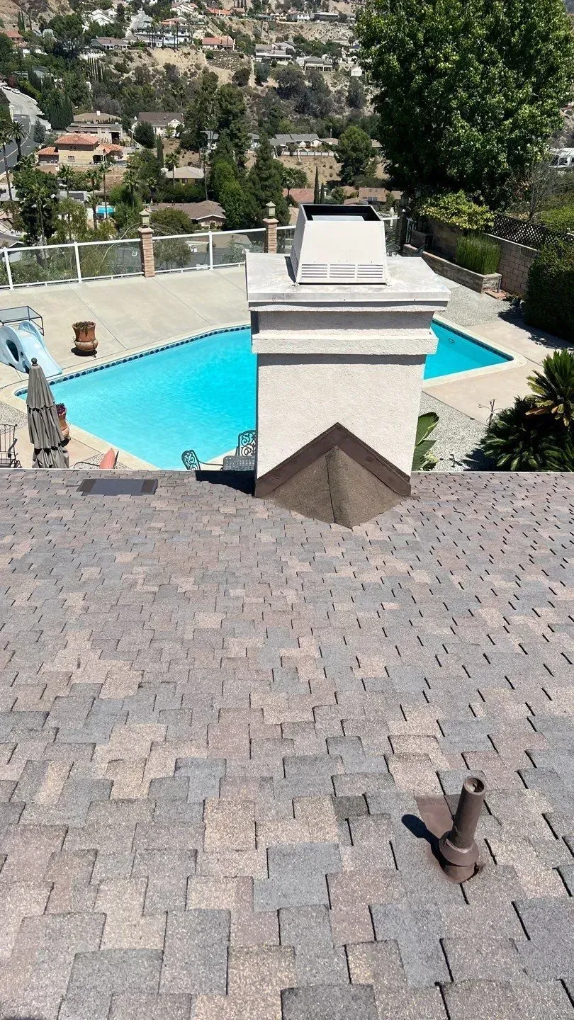 Chimney on a shingled roof with a swimming pool and hillside in the background.
