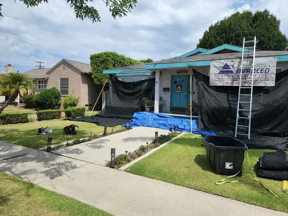 House with blue door covered in black netting for roof work. Ladder, tarp, and equipment on the lawn.