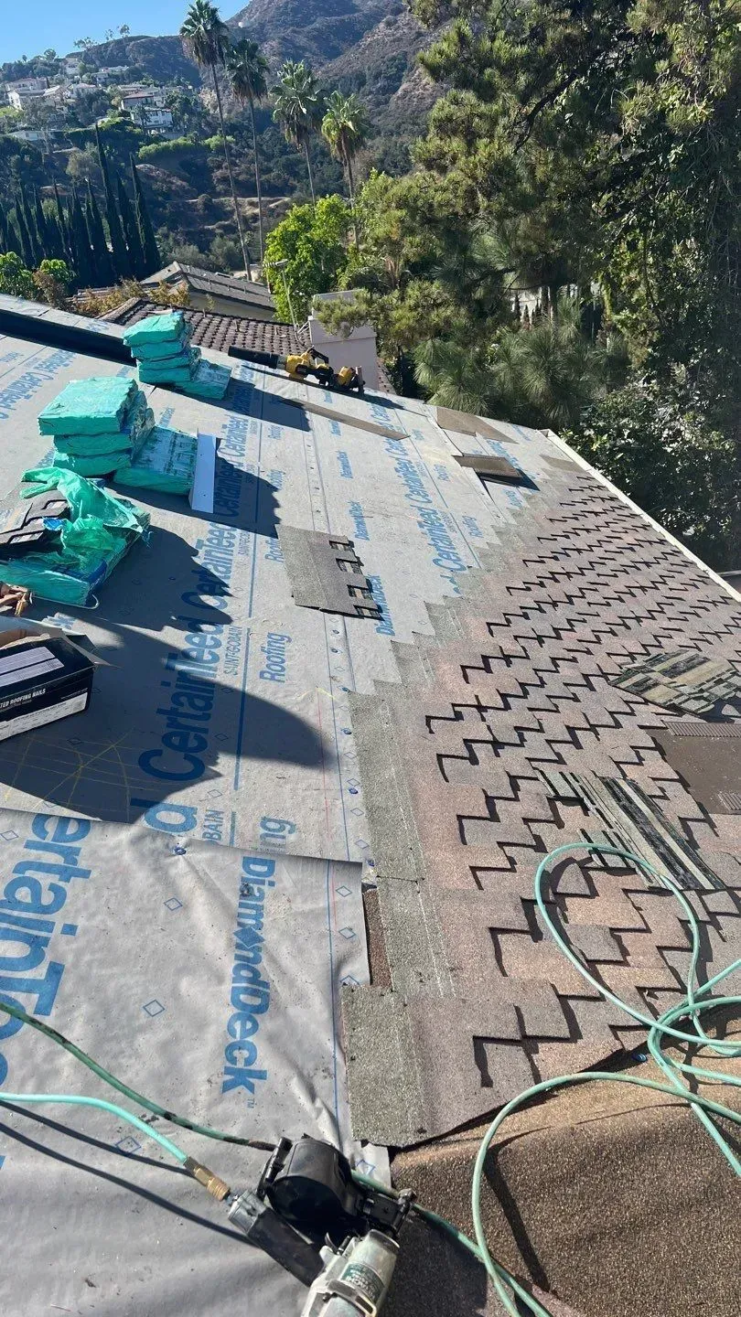 Roof partially covered with shingles, new underlayment, supplies, and mountain backdrop.
