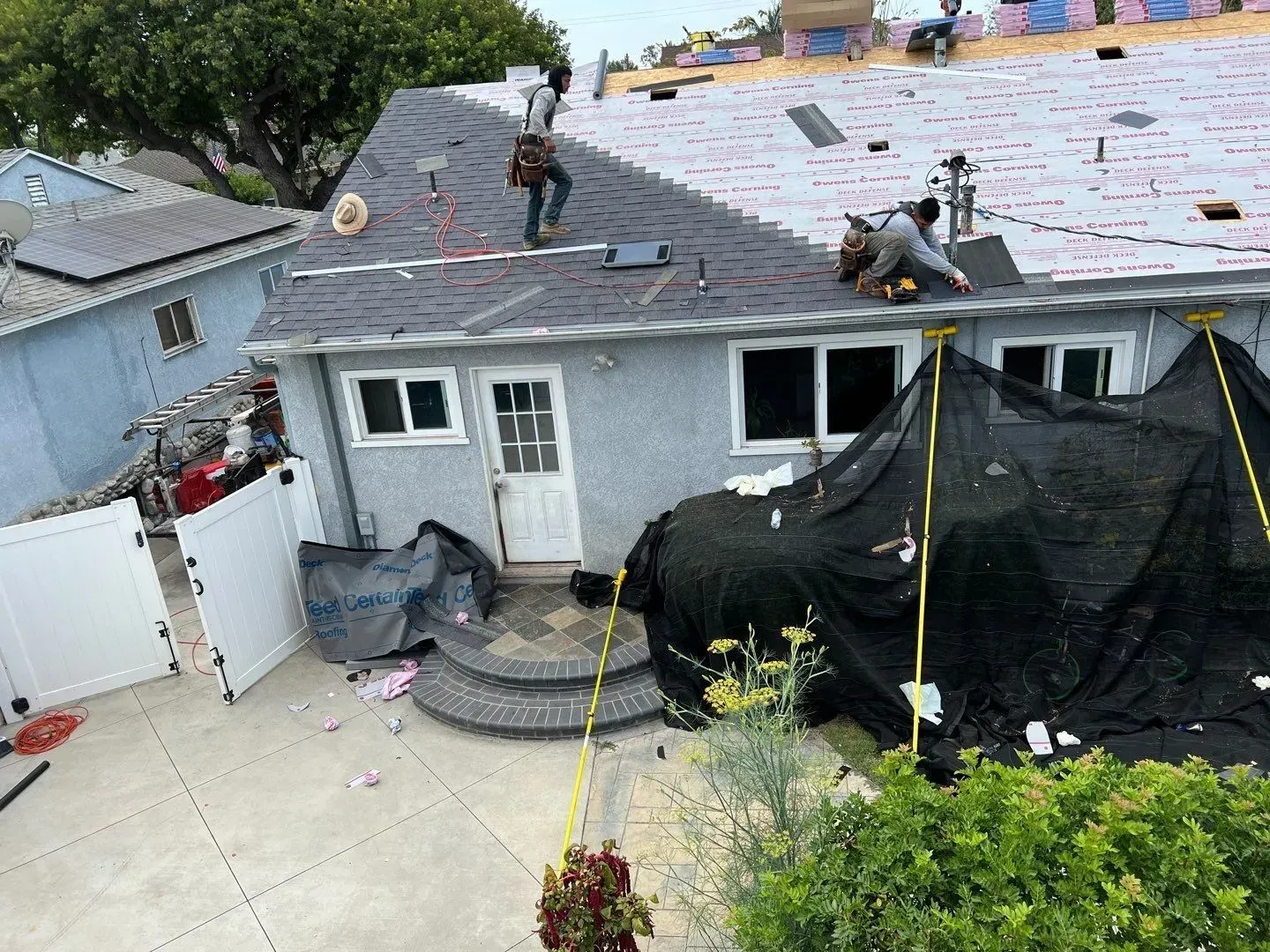 Workers installing roofing shingles on a house, with scaffolding, in a residential setting.