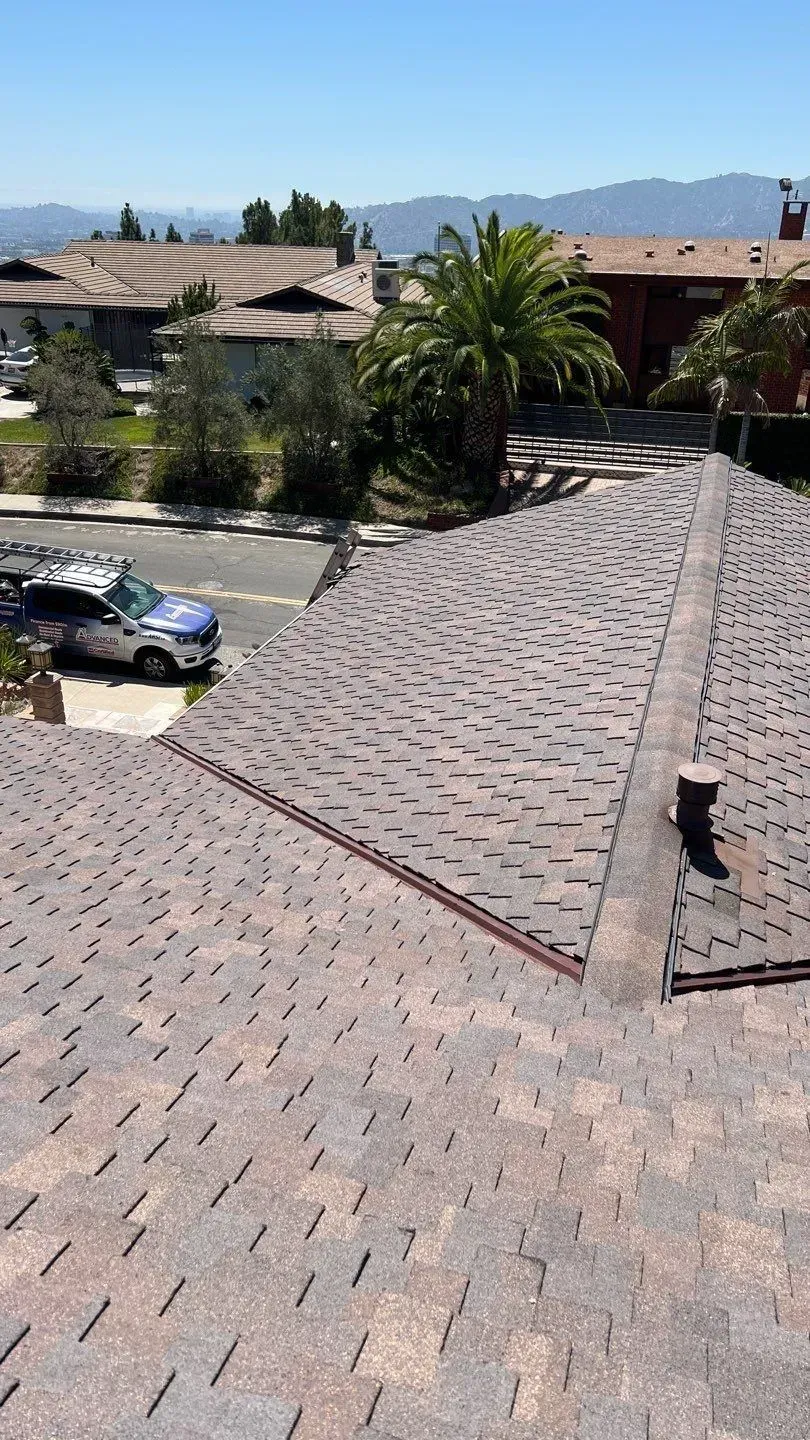 View of a brown shingle roof, street, trees, houses, and a blue & white service van on a sunny day.