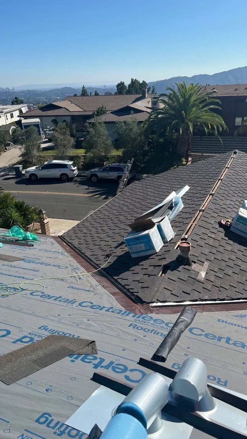 Rooftop view of a home being worked on, asphalt shingles, blue sky, houses and hills in the background.
