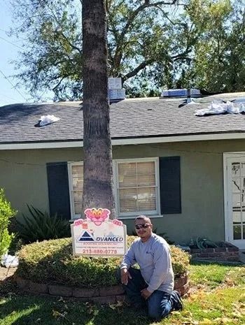 Man kneels in front of a house, a sign with a pink panther, tree, and bushes in the yard.
