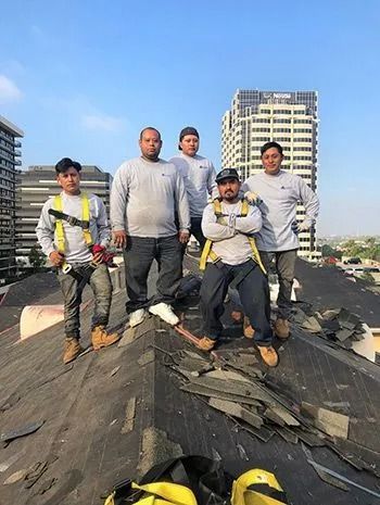 Five construction workers standing on a damaged rooftop, blue sky background.