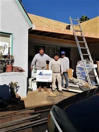 Three construction workers pose in front of a house under renovation. A ladder rests against the roof.