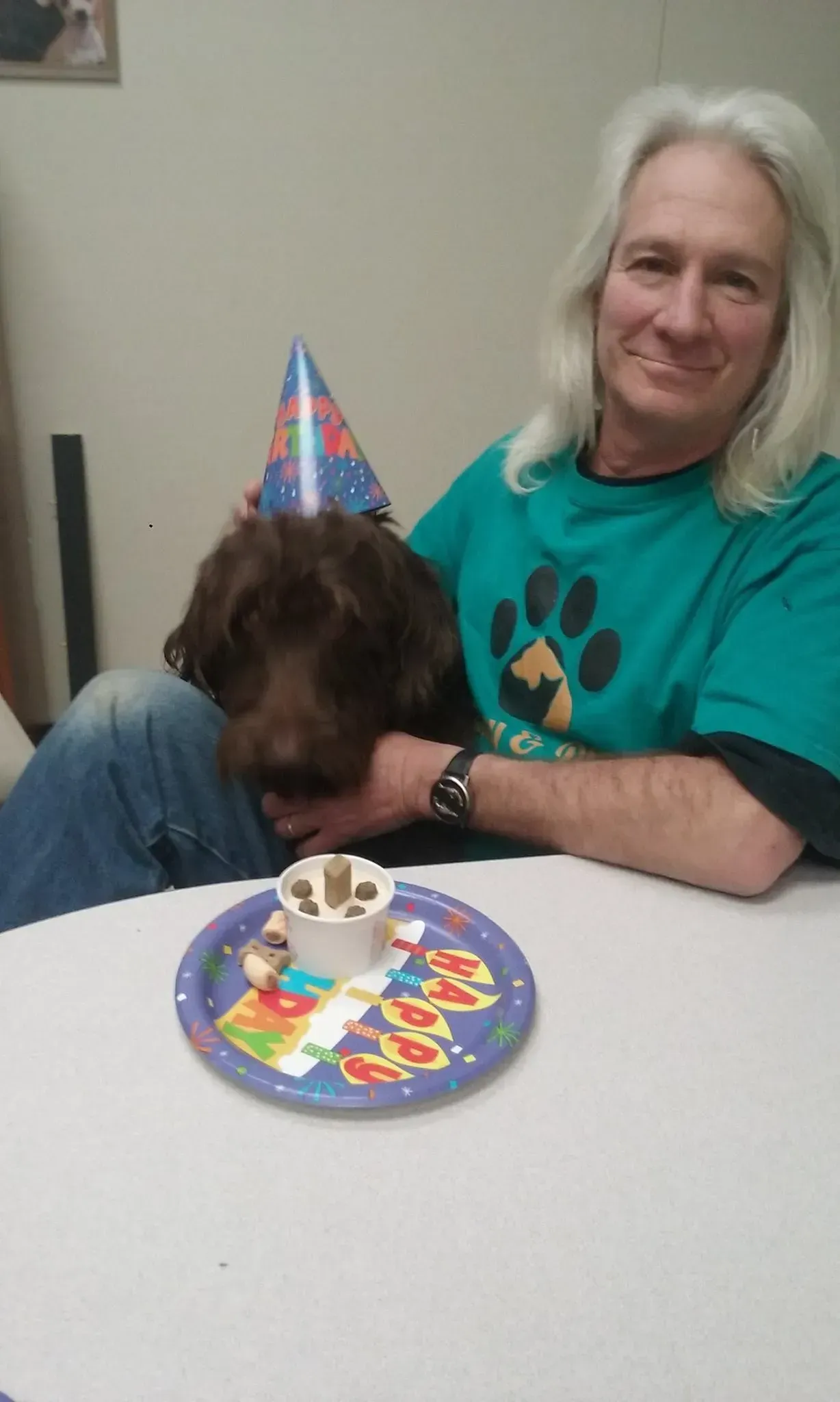 A man is sitting at a table with a dog wearing a birthday hat.