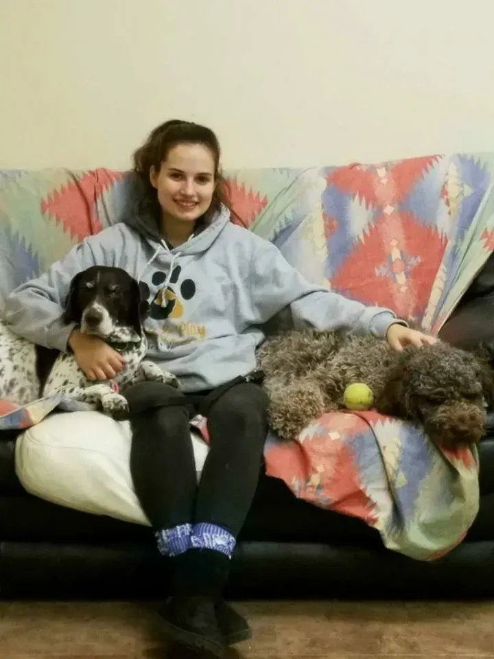 A woman is sitting on a couch with two dogs and a tennis ball