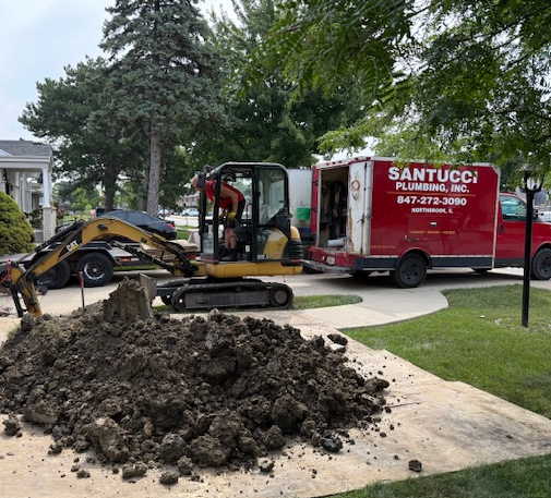 An excavator digs in a residential yard. A plumbing truck and trailer are parked nearby.