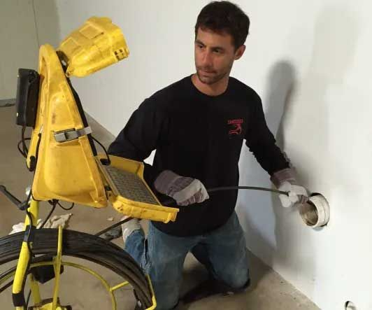 Man using drain inspection camera to examine pipe in white wall.