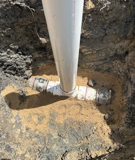 White PVC pipe and fittings in a dirt pit, with sand surrounding the pipes.