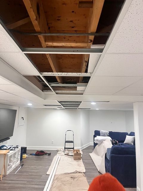 Basement ceiling renovation with exposed beams and recessed lighting. White walls, blue sofa, and tools.
